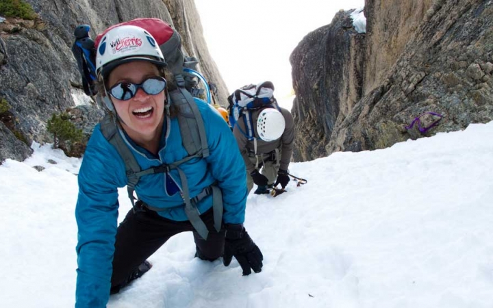 a person wearing mountaineering gear looks up and smiles at the camera while making their way up a snowy incline between two large rocks. There is another person in mountaineering gear behind them.